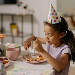 Girl at birthday party eating cake, wearing a festive hat indoors.